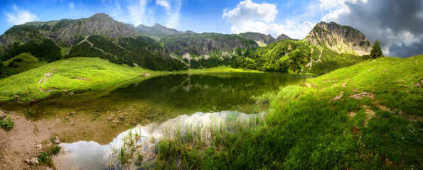 Wall Mural - Mountain lake panorama with green meadows, blue sky, dramatic clouds and reflections on the clear water in the Alps