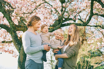 Outdoor portrait of happy young family playing in spring park under blooming magnolia tree, lovely couple with two little children having fun in sunny garden