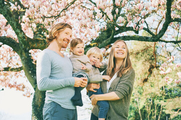 Wall Mural - Outdoor portrait of happy young family playing in spring park under blooming magnolia tree, lovely couple with two little children having fun in sunny garden