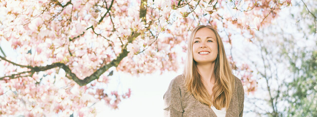 Panoramic banner background with young beautiful happy woman with blond hair, posing under blooming magnolia tree