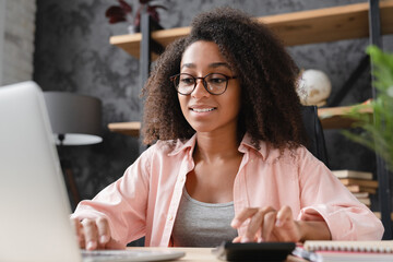 Closeup shot young african student worker businesswoman freelancer counting funds domestic bills on calculator while using laptop for e-banking, paperwork, debt and loan