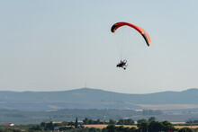 Side View Of A Paramotor Vehicle Flying In The Sky With Green And Mountains Behind