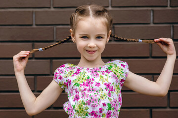 A beautiful baby girl holds herself by pigtails and smiles cutely against the background of a brick wall. Portrait of a happy child in a summer bright dress