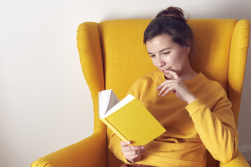 A beautiful brooding woman with a yellow book in a yellow chair. Front view.