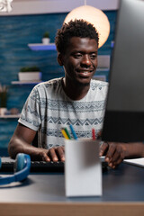 Poster - Smiling teenager entrepreneur browsing information on internet working at business project. African american businessman sitting at desk table analyzing financial graphs on computer. Remote work
