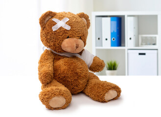 Wall Mural - medicine, healthcare and childhood concept - teddy bear toy with bandaged paw and patch on head over medical office at hospital background