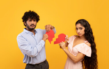 Wall Mural - End of love concept. Unhappy indian couple holding pieces of broken heart on yellow studio background