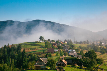 Wall Mural - Mountain panoramic landscape with trees and a village on a foggy and sunny morning.