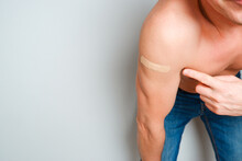 A Man Who Received A Vaccine Shows His Hand With A Band-aid. A Satisfied Man Without A Shirt On A Light Background Leaned Over The Camera And Shows A Vaccinated Hand. Place For Text.