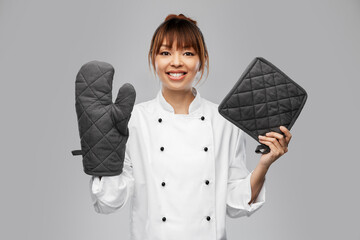 cooking, culinary and people concept - happy smiling female chef in white jacket with potholder and oven mitt over grey background