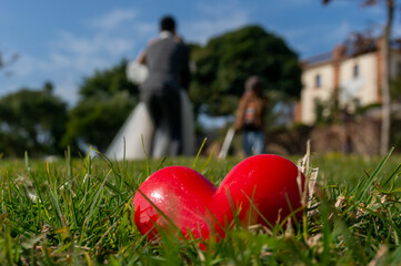 A red heart placed on a dark green lawn. The romantic backs of two lovers blurred in the distance. Ceremony at the time of marriage. Material for Valentine's Day.