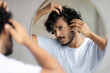 © Prostock-studio - Haircare treatment. Worried indian guy looking at flaky scalp in mirror, examining gray hair and hairloss issue