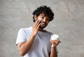 Wall Mural - Cheerful indian man applying cream on face, holding moisturizer jar in hand, smiling to camera, standing in bathroom