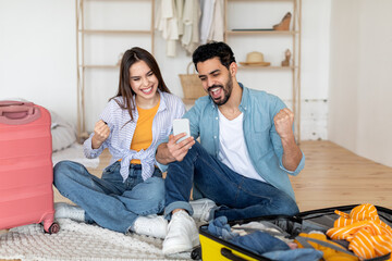 Wall Mural - Travel application. Happy emotional couple using smartphone, booking tickets, sitting near suitcases and shaking fists