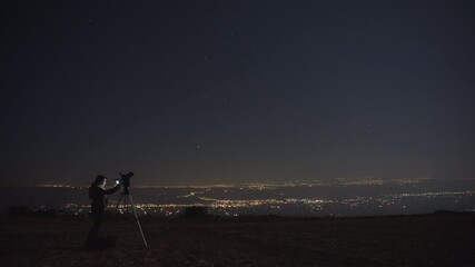 Wall Mural - Woman photographing stars with a camera and a tripod at night. City lights in the background. 