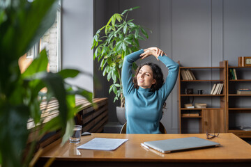 Smiling young female employee relaxing at workplace, sitting in chair stretching arms, doing gymnastic in office, looking in window with pleased face expression, feeling satisfied with work results