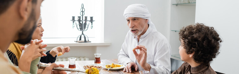 Wall Mural - mature muslim man gesturing while talking during dinner with multicultural family, banner.