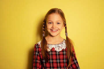 Half-length portrait of cute little girl, pupil wearing plaid dress posing isolated on yellow background. Concept of childhood, emotions, study