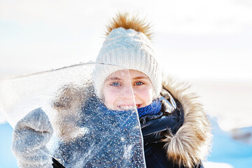 Wall Mural - Portrait of smiling little girl looks through a transparent piece of ice on a frosty winter day