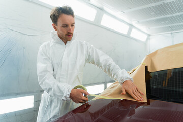 Wall Mural - Auto painter covering car body before painting or polishing with masking tape and paper. Man in a white protective suit works in a car painting booth