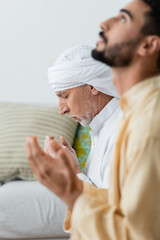 Canvas Print - Side view of mature man praying with closed eyes near blurred arabian son at home.