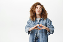 Portrait Of Smiling Curly Girl, Stylish Girlfriend Shows Heart, Love Sign And Pucker Lips, Kissing Gesture, Standing In Denim Clothes Over White Background