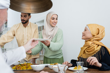 Poster - Happy muslim woman holding plate near pilaf and interracial family at home.