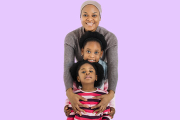 Wall Mural - Charming african muslim woman in headscarf smiling on camera while embracing her two cute daughters. Isolated over purple studio background.