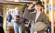 Young Stylish Woman Fitting On Classic Hat In Headwear Shop, Male Seller Holding Stacked Hats