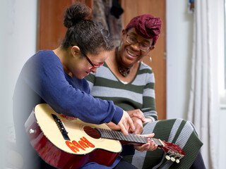 Mother assisting daughter with Down Syndrome playing guitar in bedroom