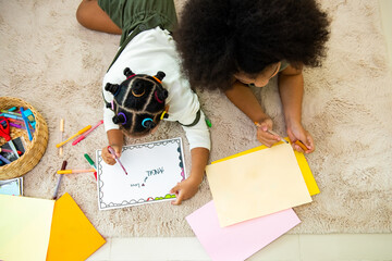 Wall Mural - Happy African cute child girl kid sibling lying on the floor using color pencil drawing and painting on the paper together. Little sister having fun indoor lifestyle leisure activity together at home.