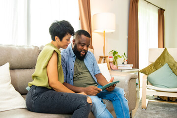 Wall Mural - African couple sitting on sofa in living room using digital tablet for online shopping with internet banking together. Happy family relax and enjoy indoor lifestyle activity with technology at home