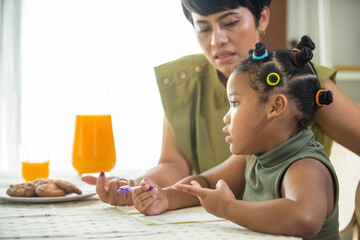 Wall Mural - African family spending time together at home. Mother helping little daughter do homework and writing on the book in living room. Cute child girl kid studying homeschooling and eating cookie with mom