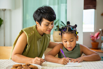 Wall Mural - African family spending time together at home. Mother helping little daughter do homework and writing on the book in living room. Cute child girl kid studying homeschooling and eating cookie with mom