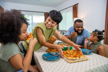 Wall Mural - African family parents and two little daughter eating fried chicken and pizza on table for dinner together. Father and mother and cute child girl kid enjoy eating and sharing a meal together at home