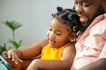 Wall Mural - Happy family African father and little daughter sit on sofa using digital tablet playing game or watching movie together. Dad and child girl kid enjoy leisure activity with wireless technology at home