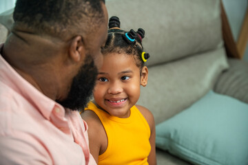 Wall Mural - Happy African family spending time together at home. Little daughter talking and playing together with father on sofa in living room. Dad and child girl kid relax and enjoy indoor lifestyle activity.