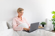 © Prostock-studio - Happy woman sitting on bed using pc