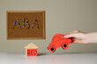 Woman holding red car near wooden house and cork board with abbreviation ABA (Applied behavior analysis) at white table, closeup