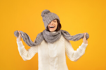 Overjoyed young lady in warm winter clothes pulling down knitted hat and playing with scarf on yellow background