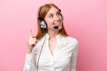 Wall Mural - Telemarketer redhead woman working with a headset isolated on pink background pointing up and surprised