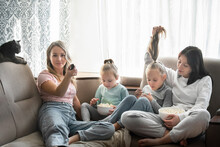 The Family Spends The Day Together At Home. Four Girls Of Different Ages Sit Together On The Couch, Eat Popcorn And Watch TV, Next To Them Is A Gray Cat.