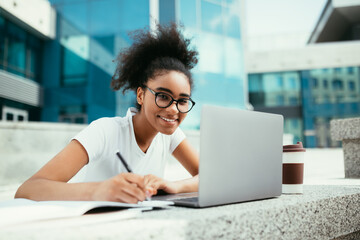 Wall Mural - Cheerful African Student Girl Taking Notes At Laptop Outdoor