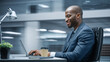 © Gorodenkoff - Modern Office: Successful Businessman Sitting at Desk Using Laptop Computer. African American Entrepreneur in Suit working with Stock Market Investing. Motion Blur Background Showing Active Work Day.