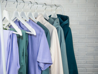 Lilac and green T-shirt, sweatpants, sweatshirt. Women's, youth clothes of different colors hang on a hanger. White brick wall.