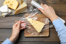 Woman Putting Plastic Food Wrap Over Block Of Cheese At Wooden Table, Top View