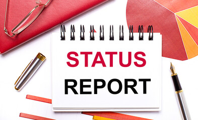 Fototapete - On the desktop is a white notebook with the text STATUS REPORT, a pen, burgundy and red tables, and gold-framed glasses. Business concept.