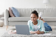 © Prostock-studio - Smiling adolescent african american girl studying, watching online lesson on laptop, lies on floor