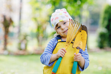 Wall Mural - Adorable little toddler girl holding garden tools in hands. Cute child learn gardening, planting and cultivating vegetables in domestic garden. Kid with shovel and rake. Ecology, organic food.