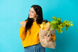 © luismolinero - Young woman holding a grocery shopping bag isolated on blue background pointing to the side to present a product